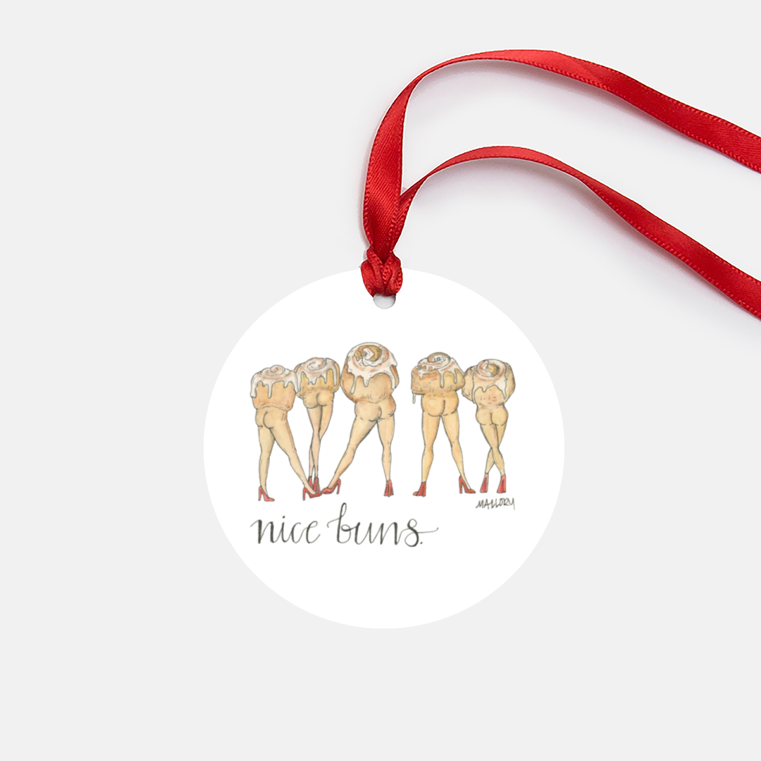 White metal Christmas tree ornament that features an illustration of cinnamon buns with legs and butts with the saying, "nice buns" on it. Ornament comes with red satin ribbon.