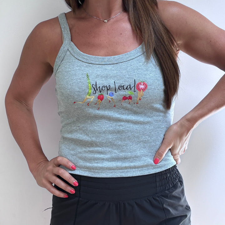 A woman wearing a light heather gray racerback tank top with a colorful 'Shop Local' graphic on the chest, paired with black athletic shorts, standing against a plain white wall.