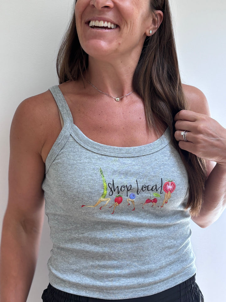 A woman wearing a light heather gray tank top with a colorful graphic that reads 'shop local' and small illustrations of fruits and vegetables, photographed against a plain light background.