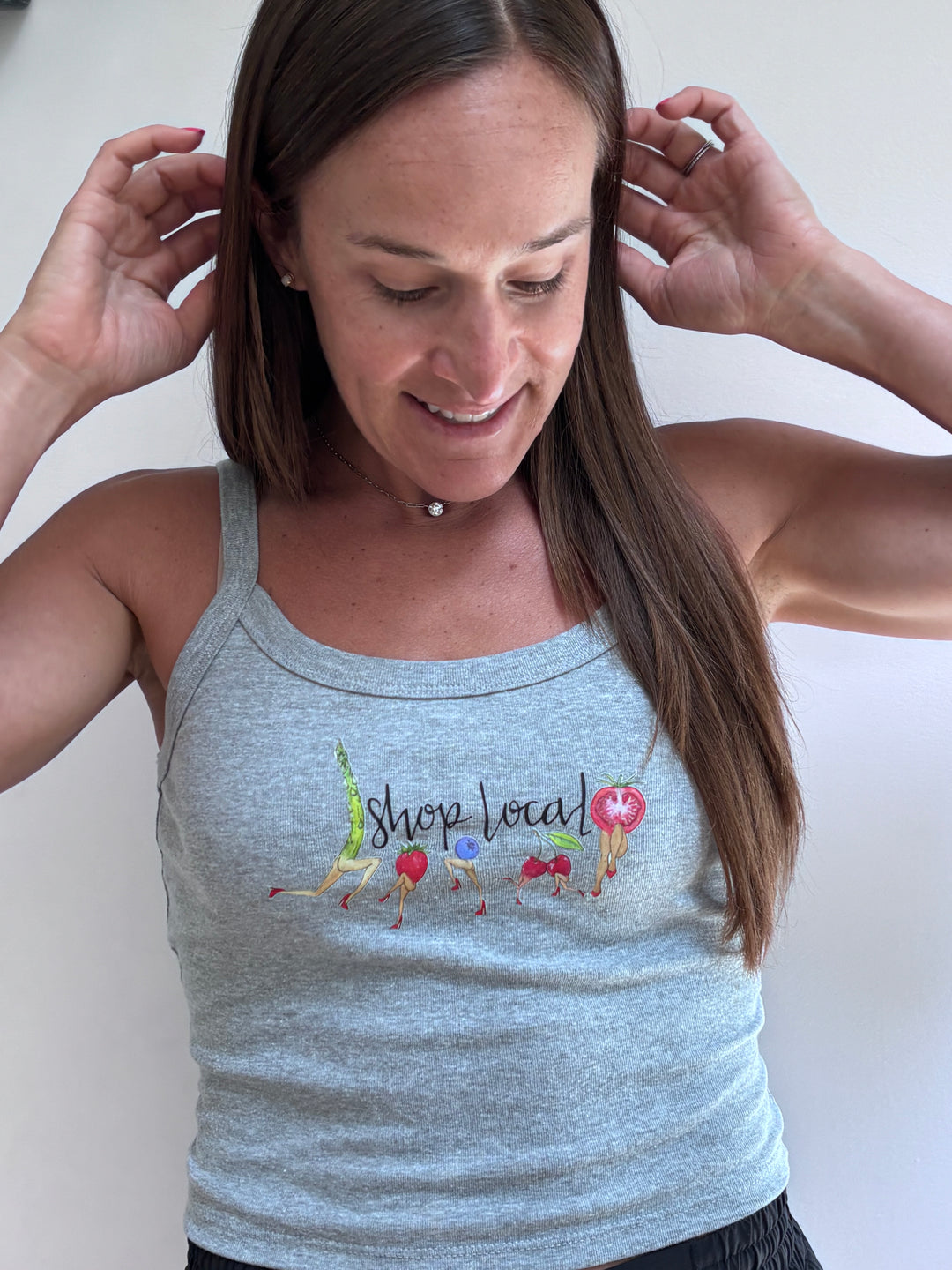 A woman wearing a light gray heather tank top with a colorful graphic that reads 'shop local' and small illustrations, photographed against a plain light background.