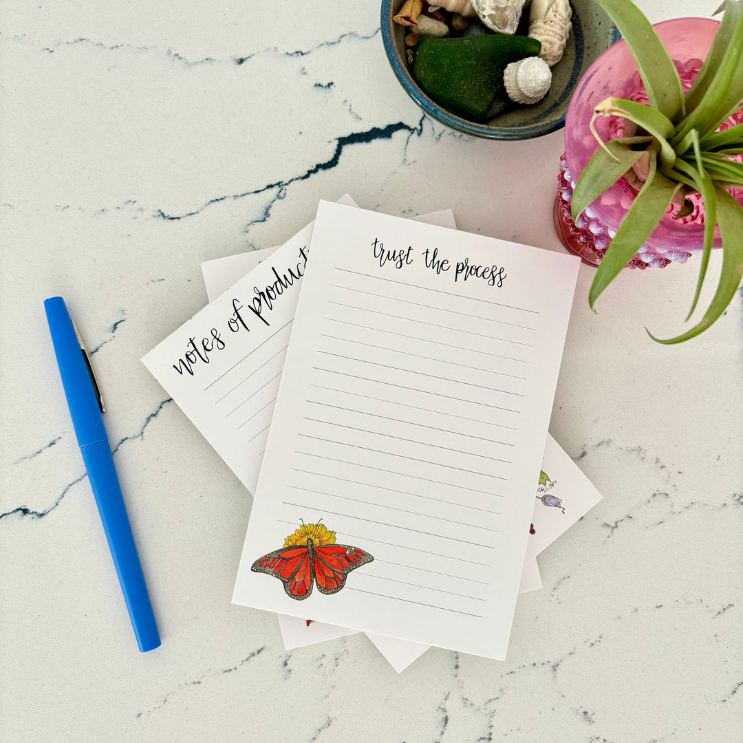 Notebook with butterfly design on a marble surface with a pen and plants.