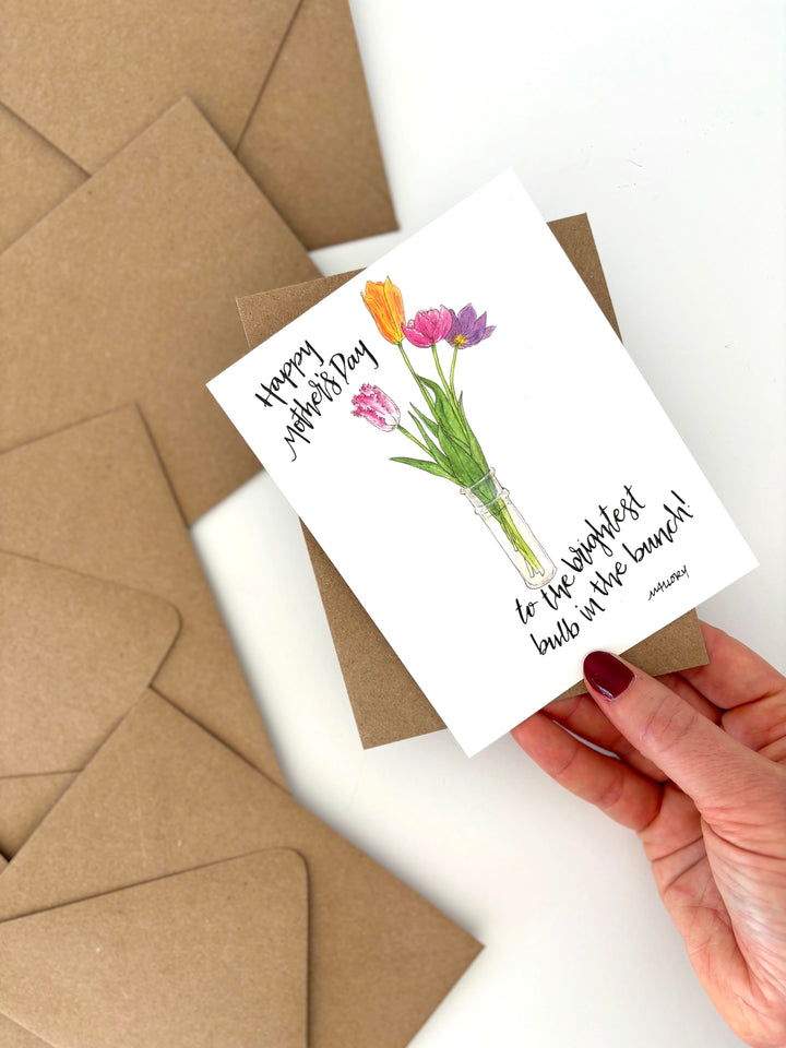 Hand holding a Mother's Day card with pink and purple tulips and envelopes on a white background. The text reads, "happy mother's day to the brightest bulb in the bunch"