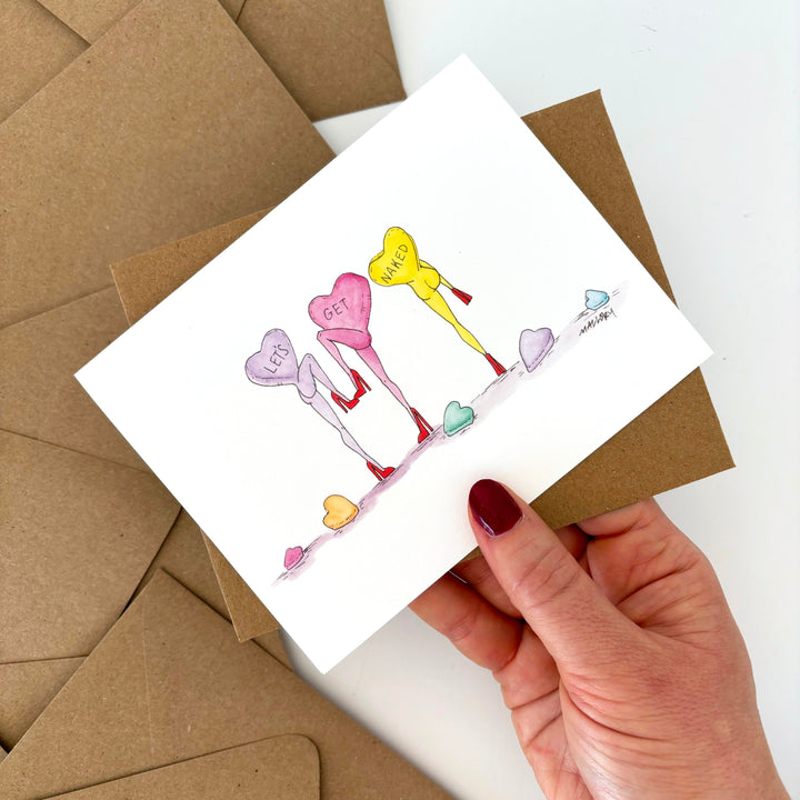 Hand holding a Valentine's Day card with colorful dancing candy hearts and envelopes on a white background. The message on the candy hearts is "let's get naked".