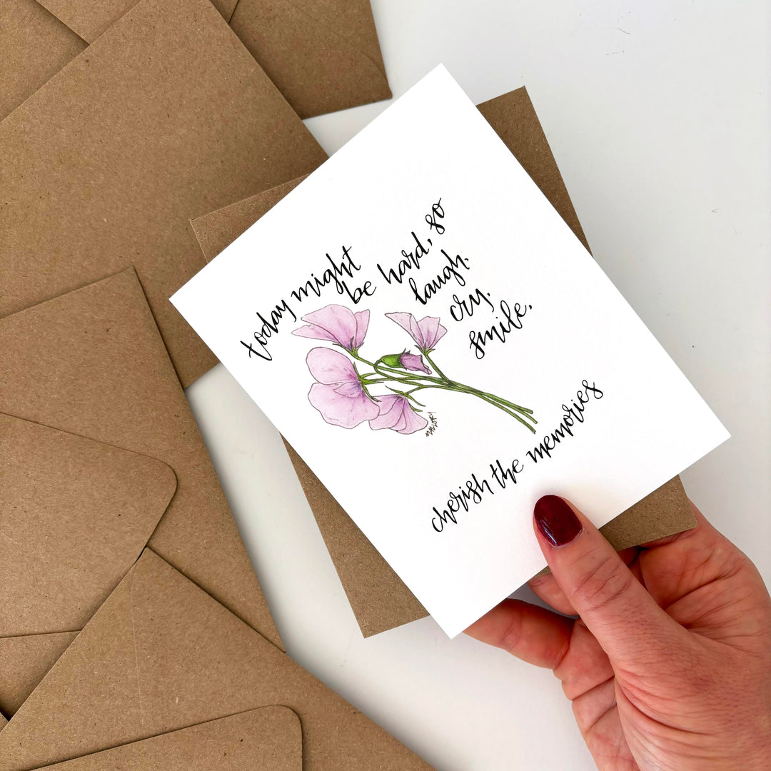 Grief card with floral design and text held by a hand, surrounded by brown envelopes. Text reads, "today might be hard, so laugh, cry, smile. cherish the memories".