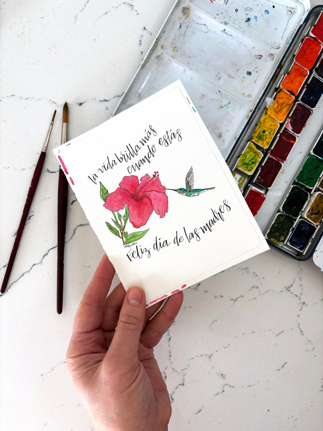 Hand holding a card with floral design and text on a marble surface with art supplies.