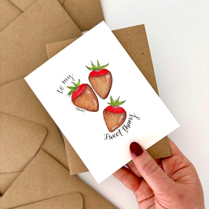 Valentine's Day card with chocolate-covered strawberry illustration held by a hand, with brown envelopes in the background.