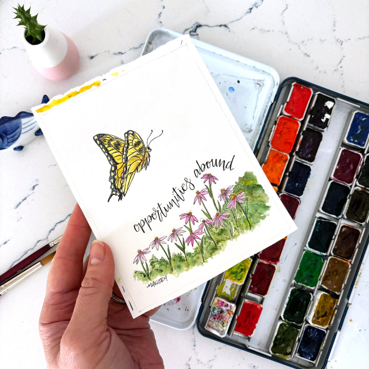Hand holding a painting with a tiger swallowtail butterfly and floral design, surrounded by watercolor paints on a marble surface.