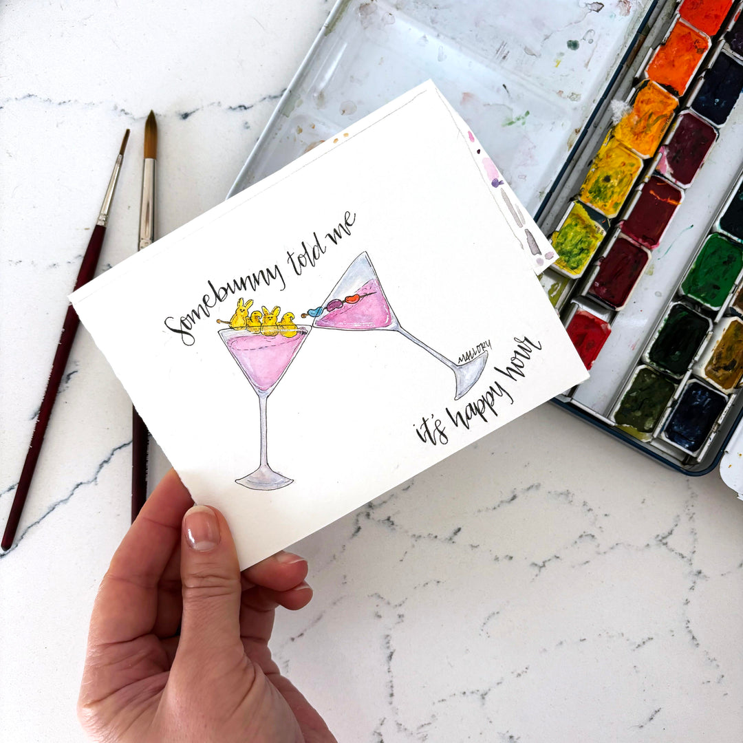 Hand holding a card with Easter cocktail illustrations and text on a marble surface with art supplies.