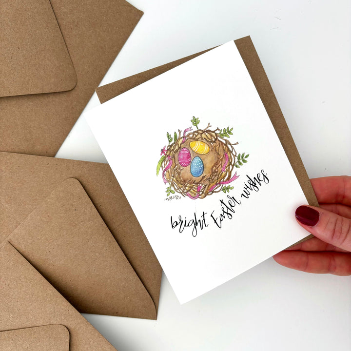 Easter card with a nest and eggs design, held by a hand with brown envelopes on a white background.