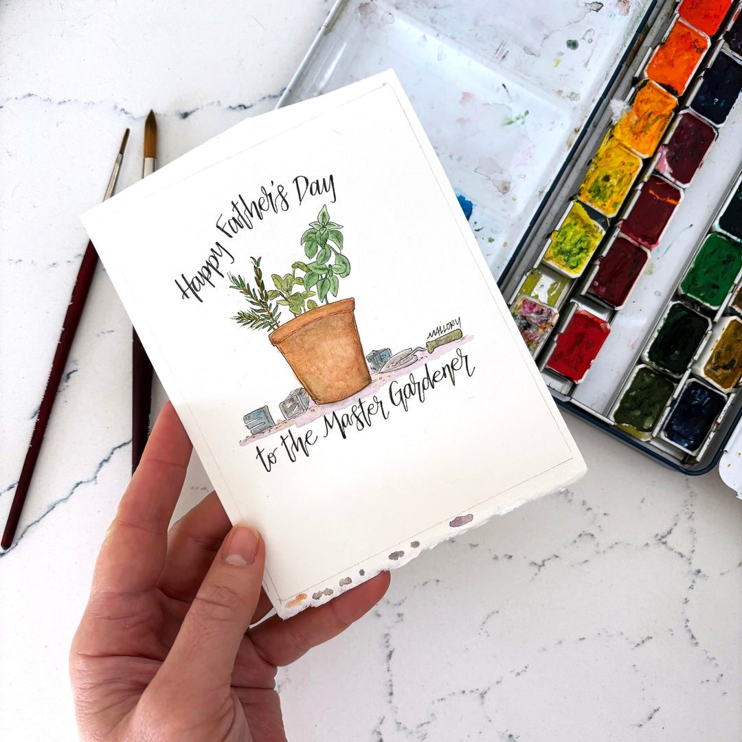 Handmade card with gardening illustration and 'Happy Father's Day' text, held by a hand on a marble surface with watercolor set.