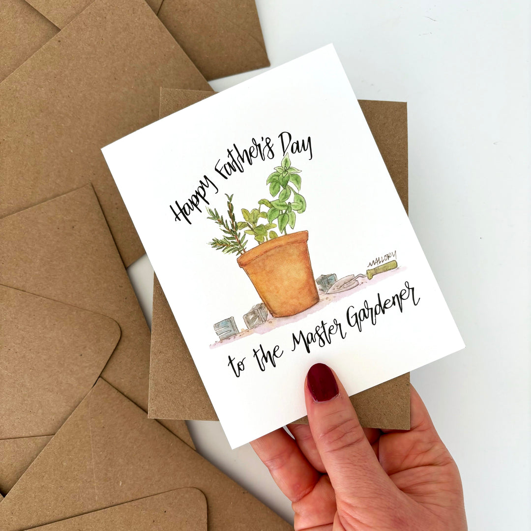 Father's Day greeting card with potted plant illustration and text, held by a hand over brown envelopes. Text reads, "happy father's day to the master gardener".