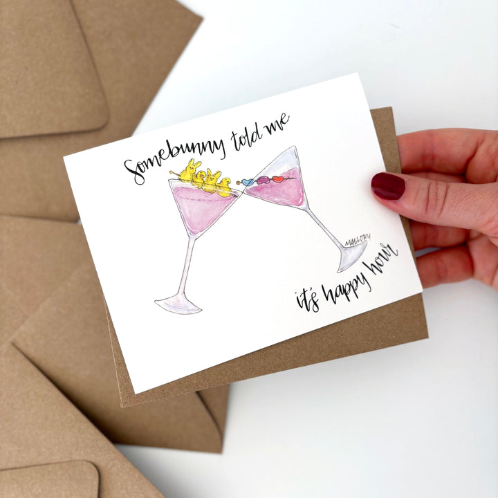 Greeting card with two cocktail glasses and text, held by a hand over brown envelopes. The Easter card has two pink martinis with Peeps and jelly bean garnishes on toothpicks.