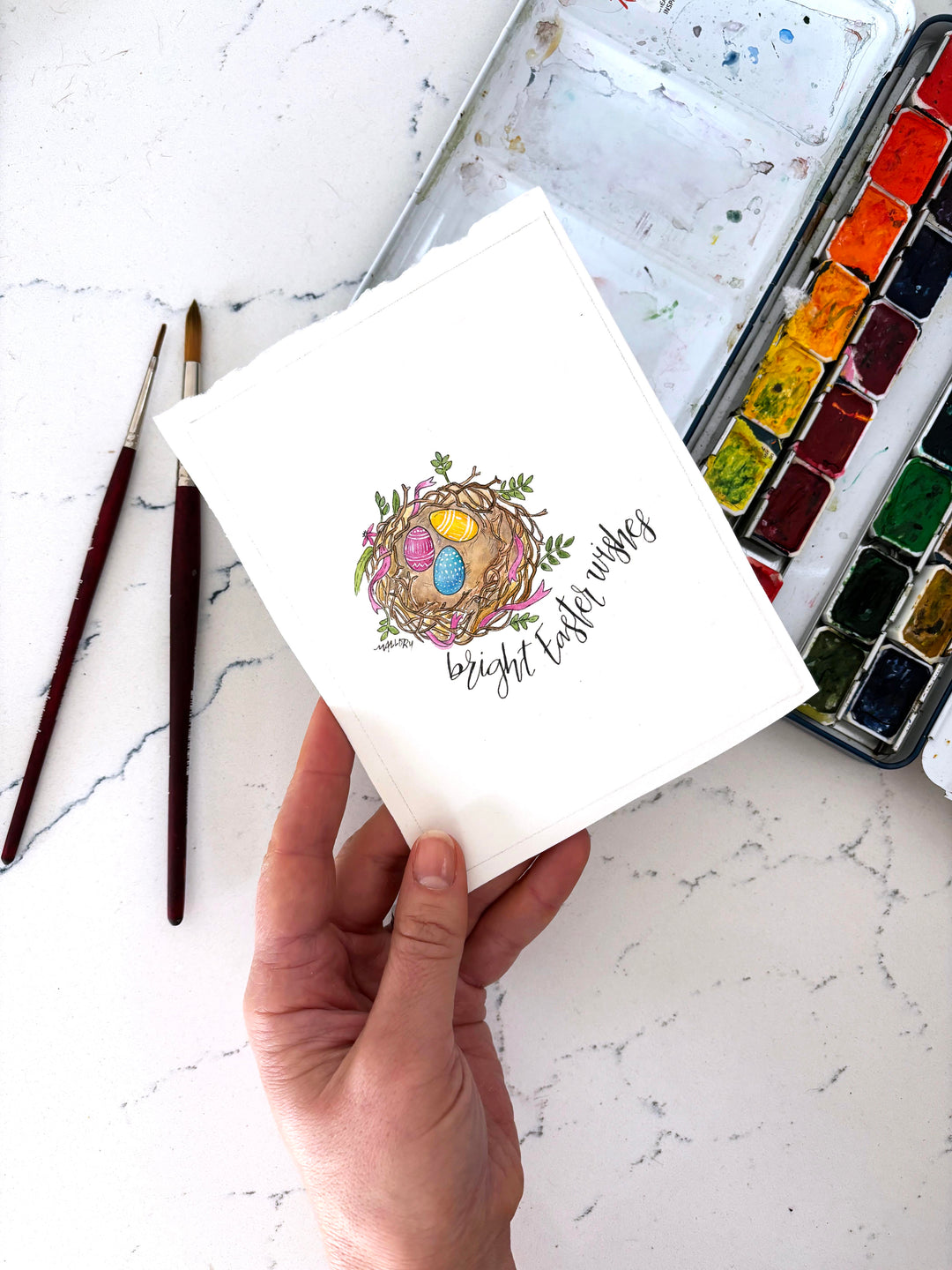 Hand holding a small watercolor card with an Easter design on a marble surface with art supplies.