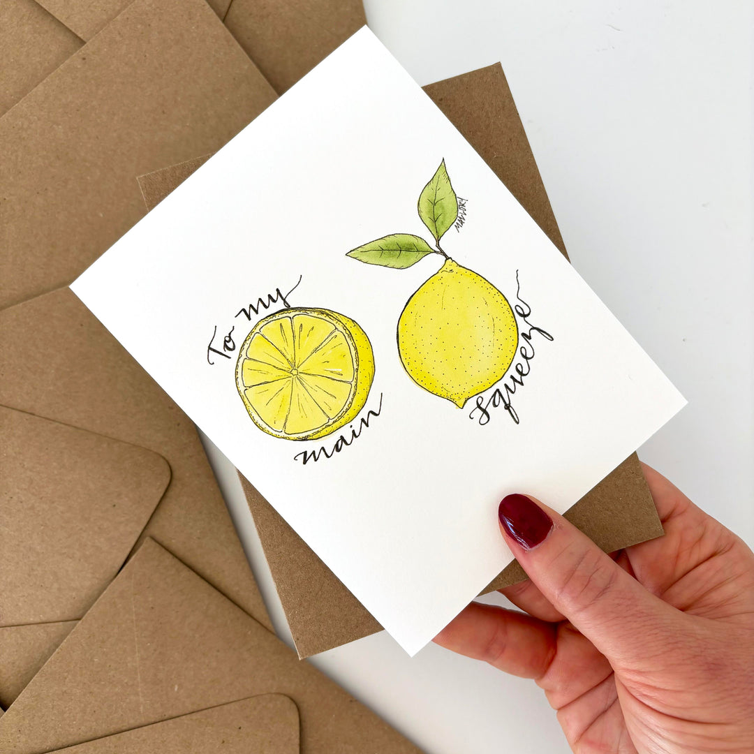 Valentine's Day card with lemon illustration held by a hand over brown envelopes. Text reads, "to my main squeeze".