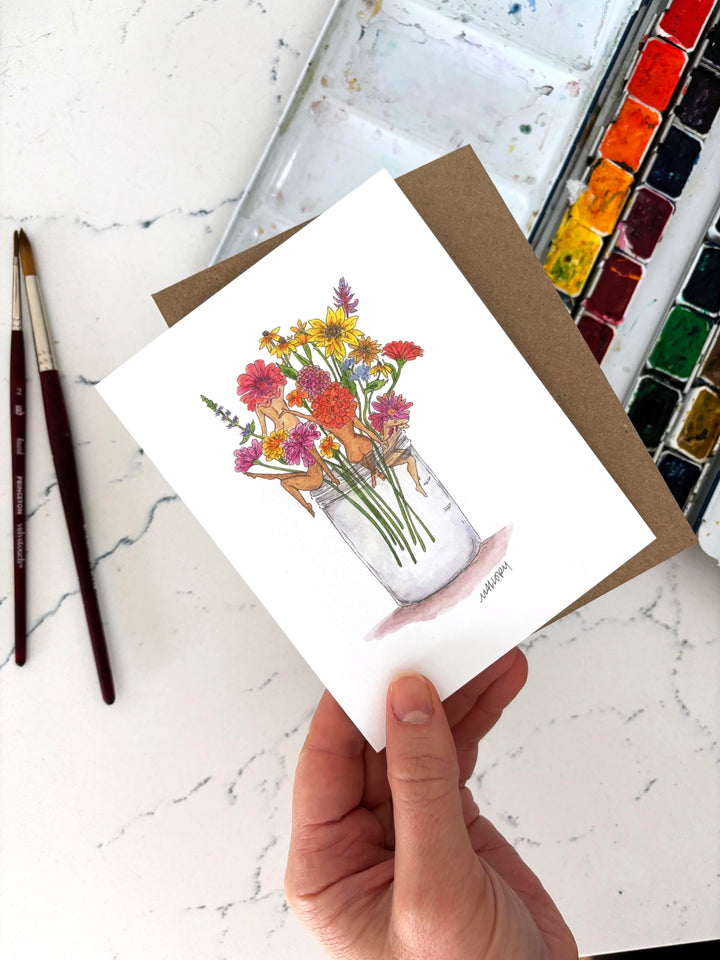 Hand holding a card with a floral design with nude women on a marble surface with art supplies.