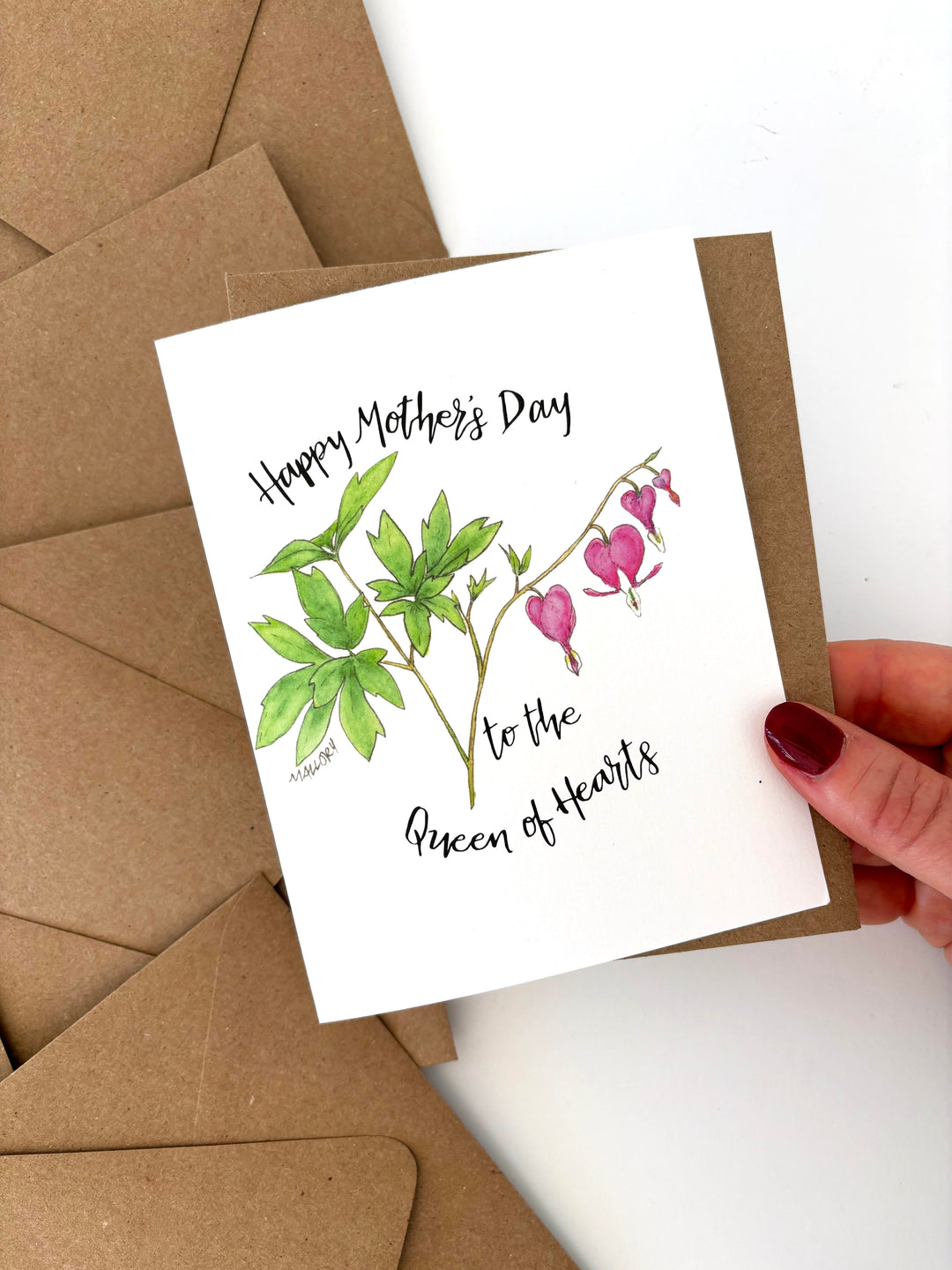 Card with floral design and text held by a hand, surrounded by brown envelopes. Text reads, "happy mother's day to the queen of hearts".