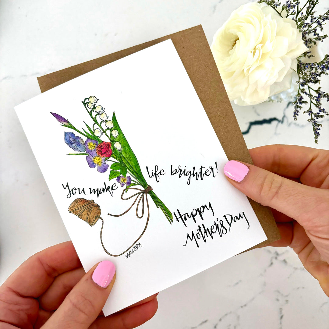 Hands holding a mother's day card with an illustration of a bouquet of flowers held together by twine. The words "you make life brighter happy mother's day" are written around the design.