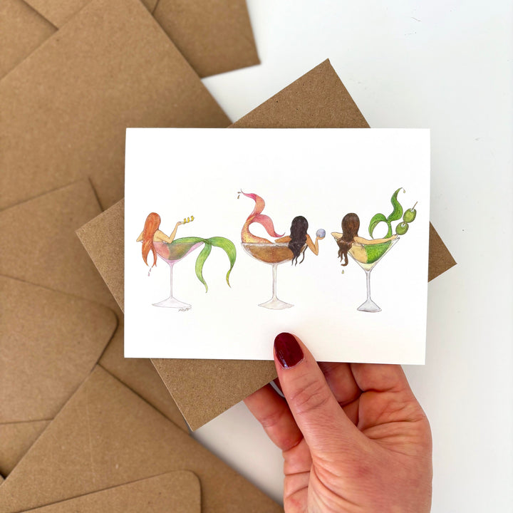Card with mermaid-themed cocktails held by a hand over brown envelopes