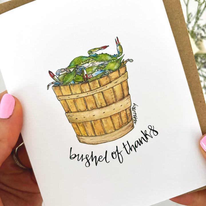 hand holding a thank you card with an illustration of blue crabs in a basket. The words "bushel of thanks" are written around the design.