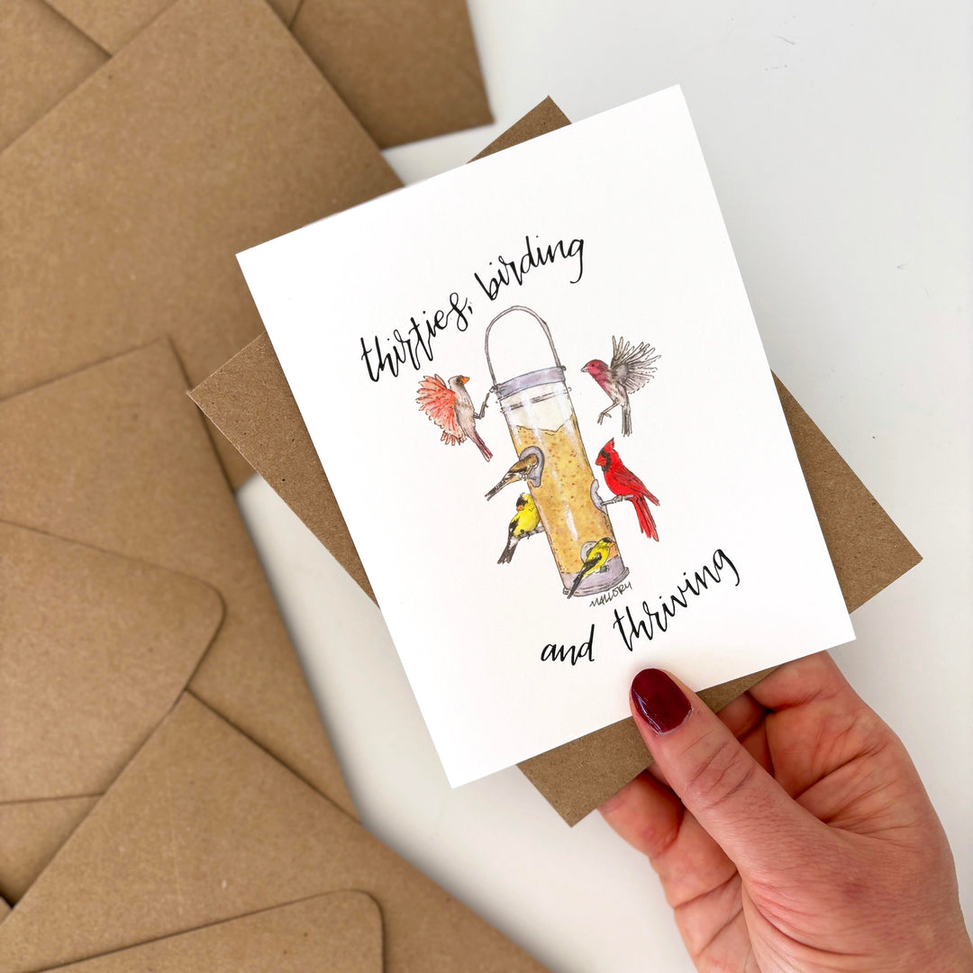 Birthday card with bird feeder illustration and text, held over brown envelopes. Text reads, "thirties, birding and thriving"