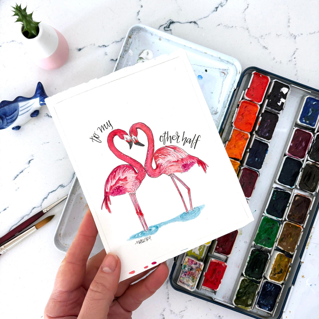 Hand holding a card with flamingo illustration and watercolor set on marble surface. The flamingos necks are in the shape of a heart for Valentine's Day.