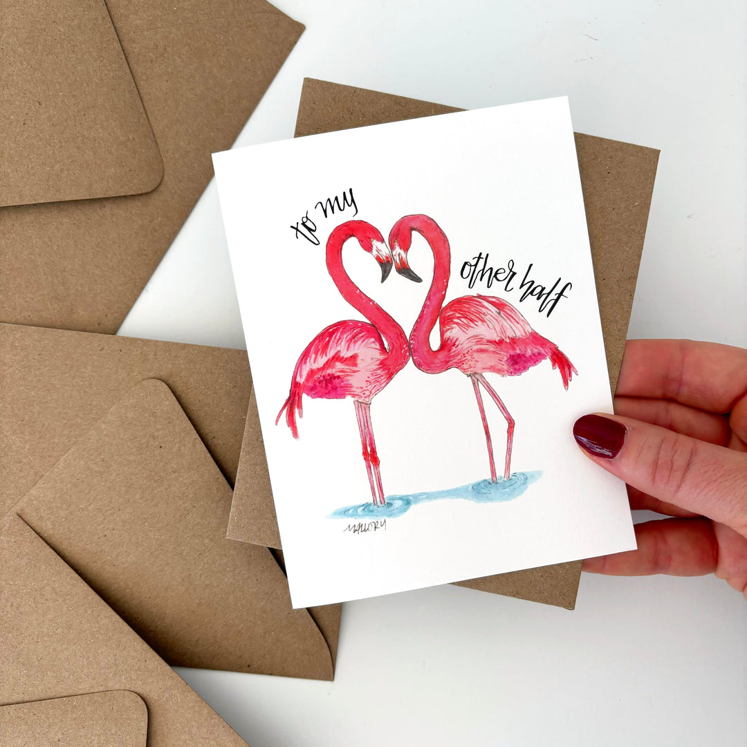 Valentine's Day card with flamingo design held by a hand, surrounded by brown envelopes on a white background. Text reads, "to my other half".