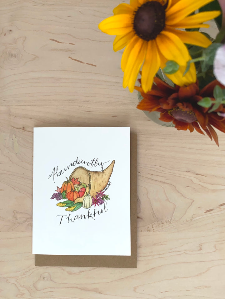 Thanksgiving card with a cornucopia filled with fresh pumpkins and vegetables in orange, green purple and natural colors. The words, "Abundantly Thankful" are depicted on the Thanksgiving card.