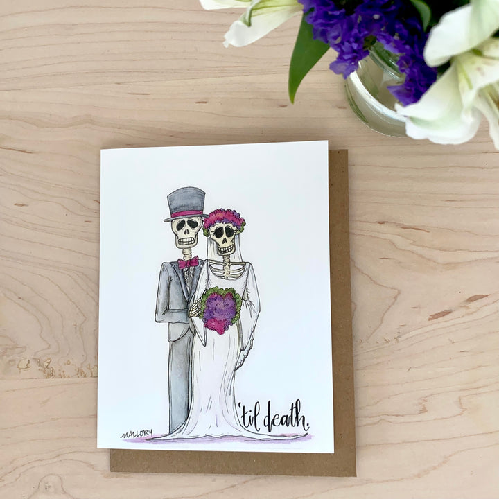 Lifestyle image of a handmade wedding or anniversary card with skeletons and flowers. Pictured with purple and white flowers.