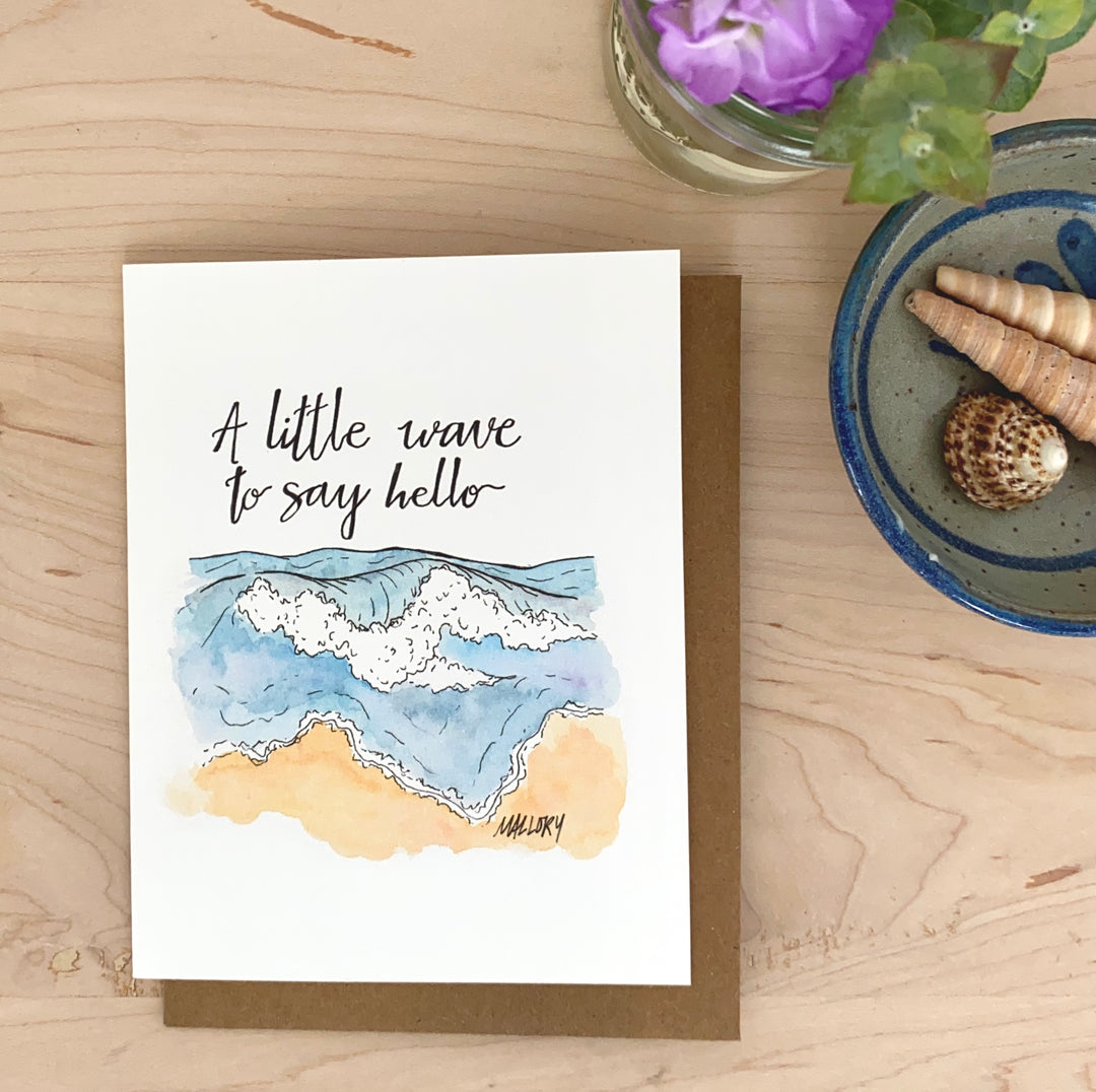 Image of a handmade greeting card with an ocean wave and beach, with the phrase "a little wave to say hello" hand-lettered on the image.