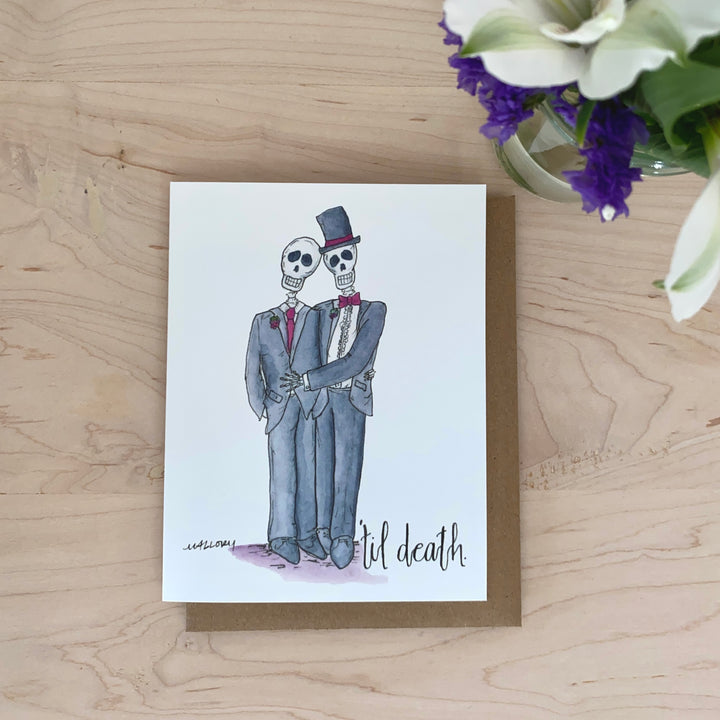 Lifestyle photo of a same-sex male couple as skeletons, with the phrase "'til death" on the bottom. Pictured with purple and white flowers.