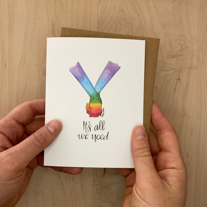 Image of a pride card to show support for LGBTQ+. Hand-drawn card shows two holding hands that are painted with the pride rainbow with the words, "It's all we need" lettered below.