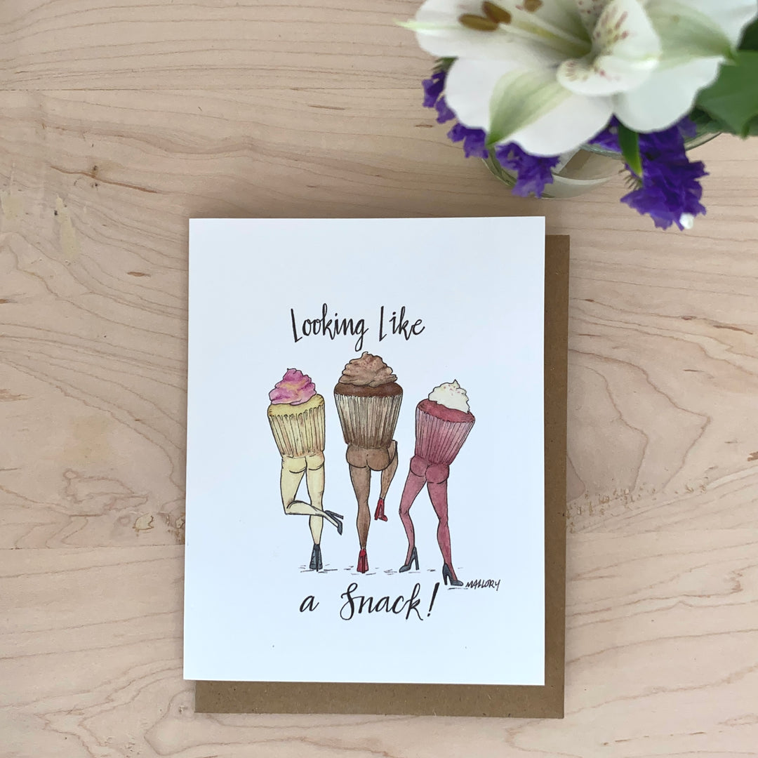 Image of greeting card on kraft recycled envelope. Card is illustrated with a vanilla cupcake, a chocolate cupcake and a red velvet cupcake with butts. The card reads, "Looking like a snack!".