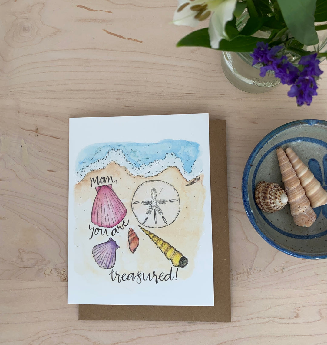 Image of a Mother's Day card with the words, "Mom, you are treasured". On the card is a blue ocean wave crashing onto a beach with pink seashells, purple and yellow seashells and a sand dollar in the sand.