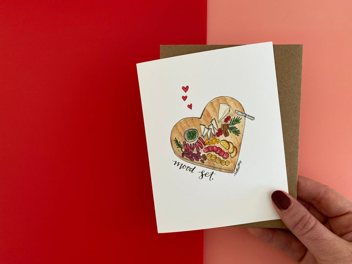 Valentine's Day card with a heart-shaped wooden charcuterie board. The charcuterie board is covered with meats and cheeses, with small red hearts floating above the board. The words, "mood set" are handlettered along the cutting board.