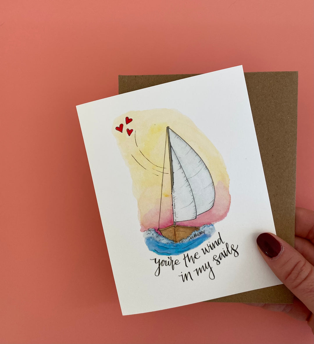 Valentine's Day card with a sailboat against a pink and yellow sunset. Little red hearts float above the sailboat, with the words "you're the wind in my sails" hand-lettered below the white wooden sailboat. The Valentine's Day card is shown against a blush pink background.