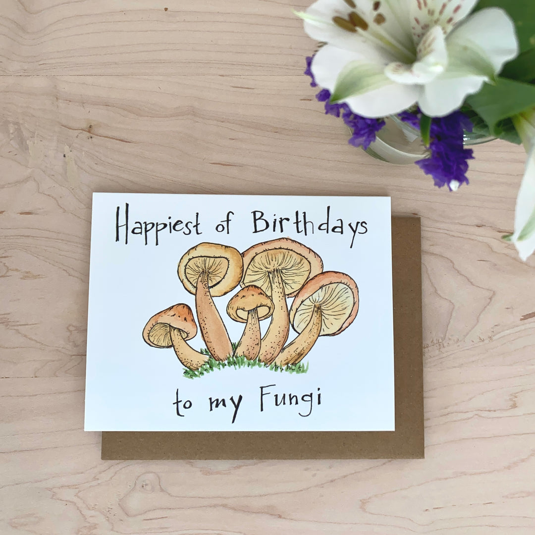 Image of a birthday card with mushrooms in the grass. The words "Happiest of birthdays to my fungi" are hand-lettered on the card.