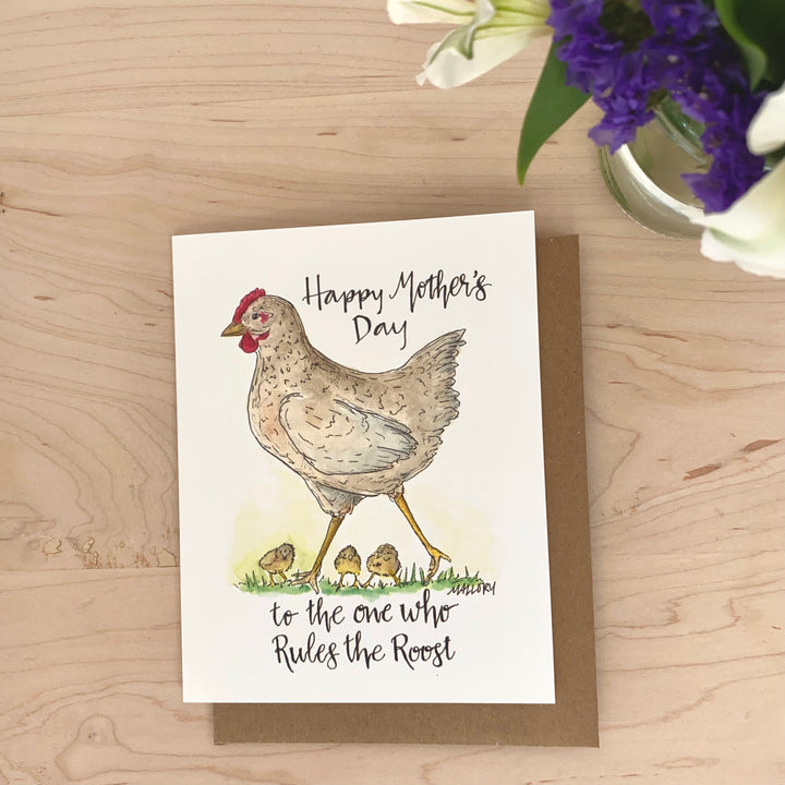 Image of a Mother's Day card with the words, "Happy Mother's Day to the one who rules the roost". On the card is a gray mother hen with little chicks running around her feet on green grass.