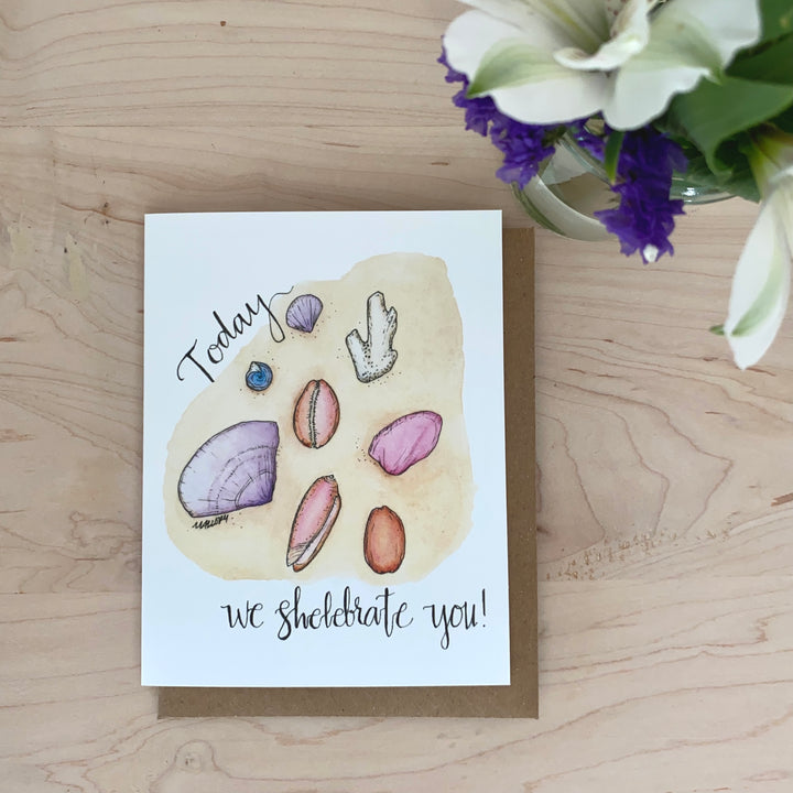 Image of a birthday card with a light sandy beach with pink, purple and ivory seashells. The words, "Today we shelebrate you!" are hand-lettered on the card.