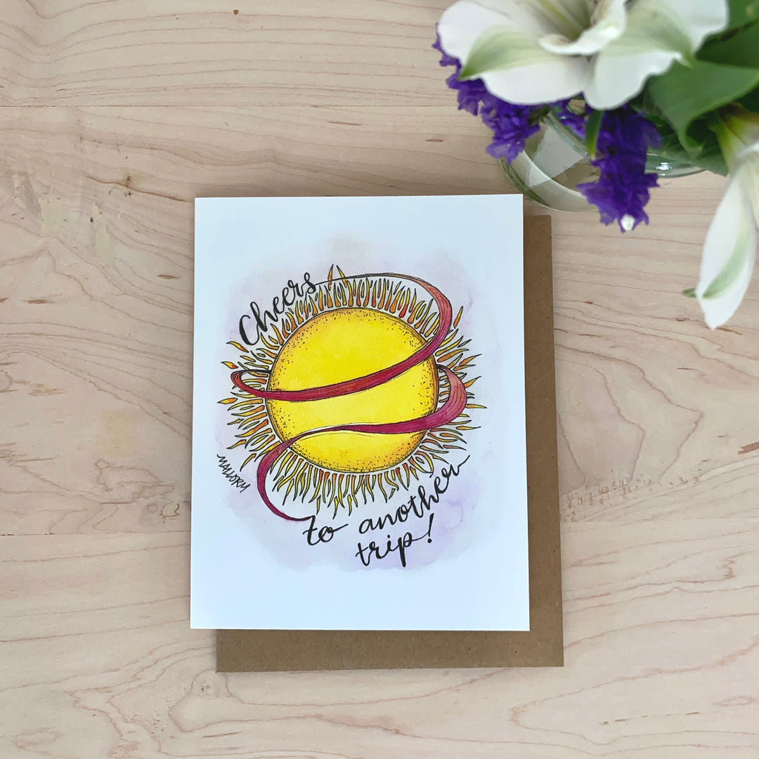 Image of a birthday card with a bright yellow sun and the words "cheers to another trip" are hand-lettered around the sun.